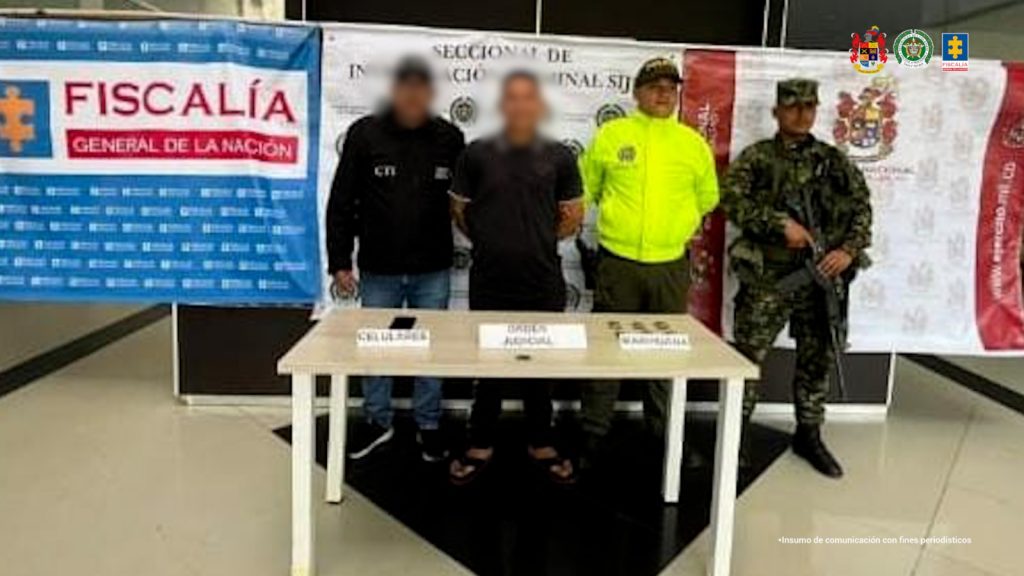 Se visualiza al capturado junto a un investigador del CTI y uniformados de la Policía y Ejército Nacional. Detrás los banners que identifican a la Fiscalía General de la Nación, Policía y Ejército Nacional.