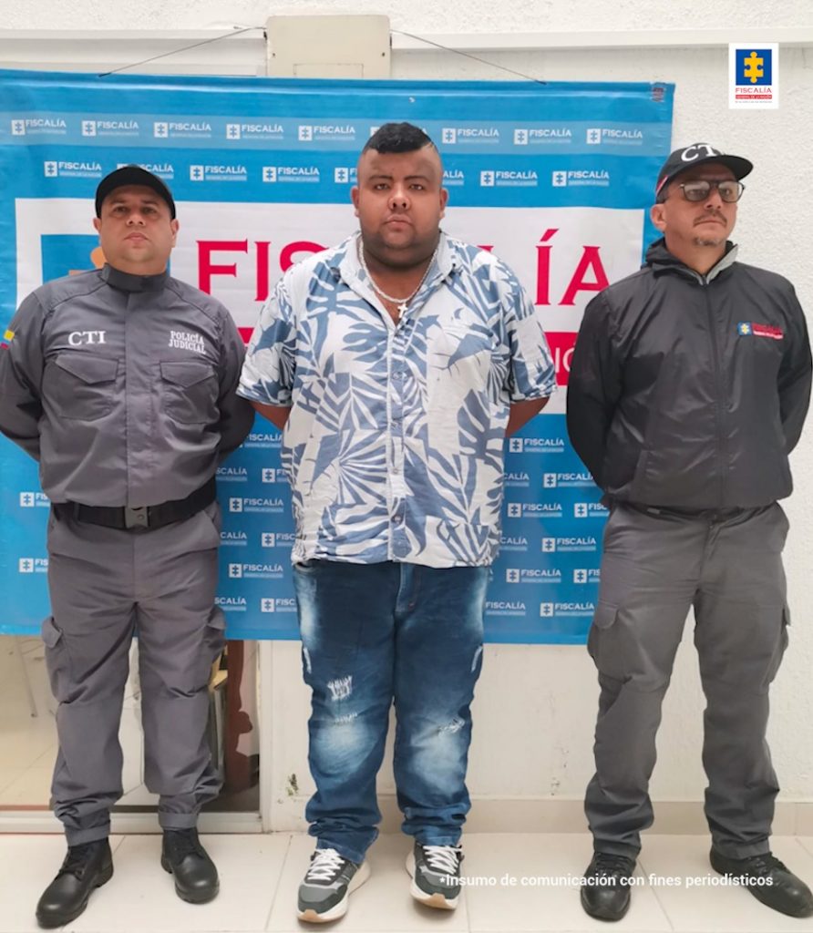 En la foto aparece, de pie y con los brazos atrás, Jhon Jairo Muñoz Salinas. Viste camisa blanca con estampados azules, jean azul, zapatos azules con blanco y usa una cadena con un dije en forma de cruz. A lado y lado de él hay servidores del CTI de la Fiscalía y detrás de ellos un pendón institucional de la Fiscalía.