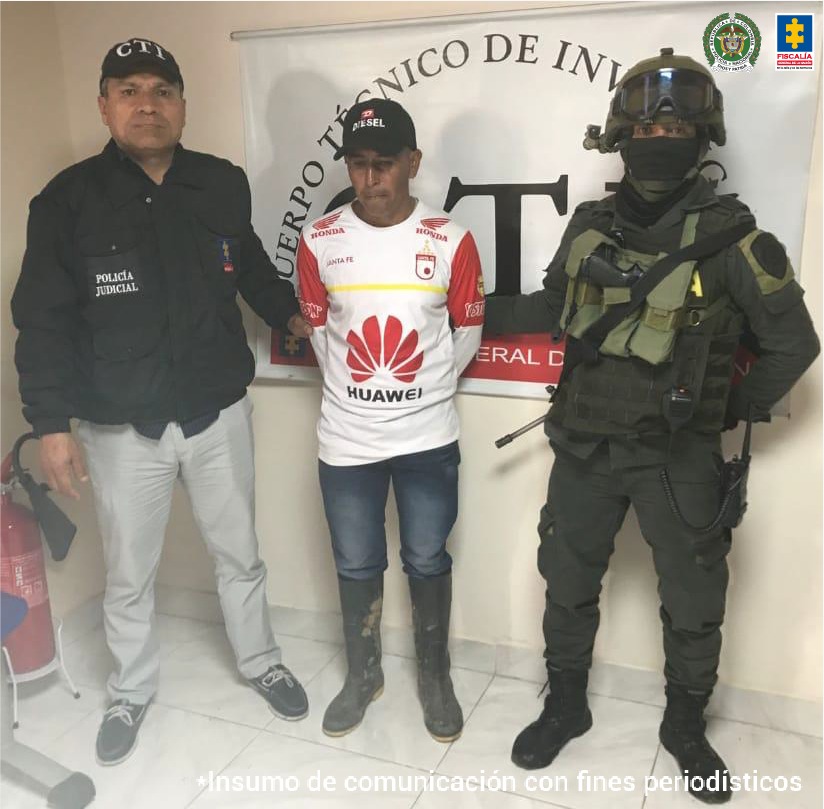 En la foto aparece Jhon Jairo Zambrano Guacheta con los brazos atrás, vestido con camiseta blanca y estampados en rojo, gorra, jean azul y botas pantaneras. Al lado derecho de él hay un servidor del CTI y a su lado izquierdo un uniformado de la Policía Nacional. Detrás de ellos hay un pendón institucional del CTI de la Fiscalía.