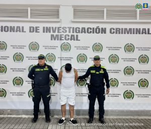 En la fotografía se aprecia de pie y con los brazos atrás a Jhon Janner Flórez Delgado. Viste camiseta esqueleto de color blanco; bermuda de color blanco y zapatillas negras con blanco. A sus extremos hay uniformados de la Policía Nacional, Detrás de ellos hay un pendón institucional de la Policía.