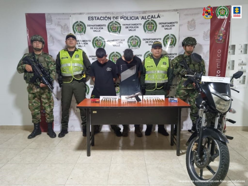 En la foto aparecen Jhon Sebastián Chávez y Luis Alfonso Cerquera, con la cabeza agachada y los brazos atrás; vestidos con camisetas oscuras. A lado y lado de ellos hay uniformados del Ejército y la Policía Nacional. Delante de ellos hay una mesa y sobre ella hay un arma de fuego, munición y un teléfono celular. Al lado izquierdo de la mesa hay una motocicleta. Detrás de ellos hay dos pendones institucionales del Ejército y la Policía.