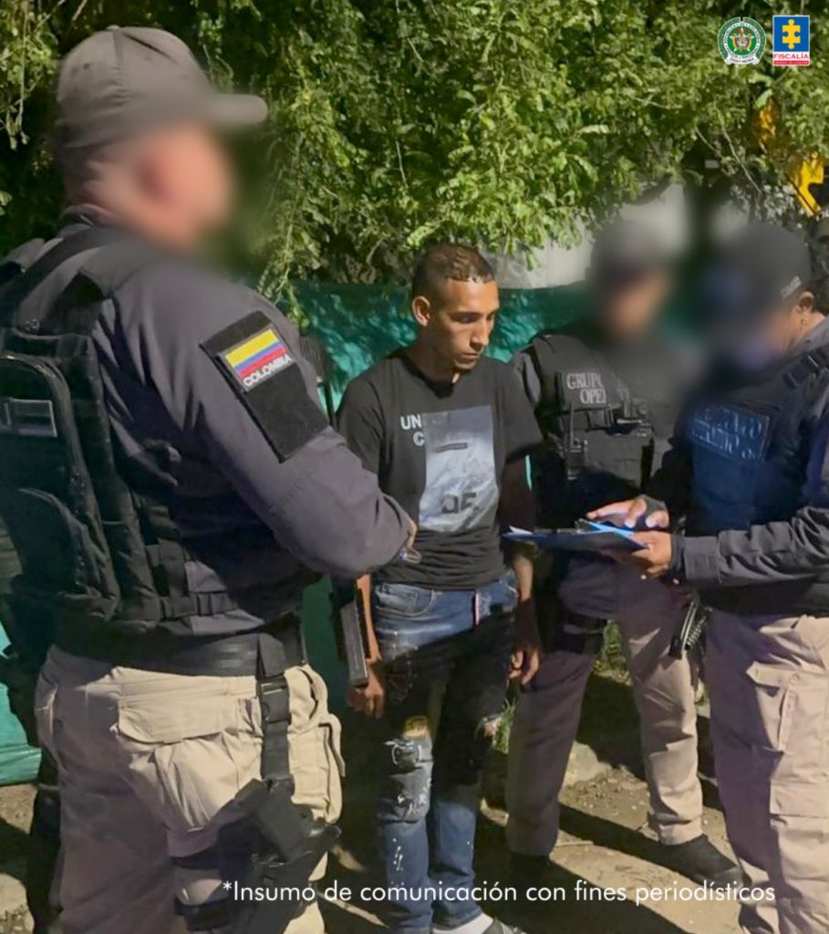 En la fotografía aparece de pie Jhon Sebastián Naranjo Mosquera. Viste con camiseta de color negro, manga corta y con estampado gris en el pecho; así como jean azul desgastado. A sus extremos hay servidores del CTI de la Fiscalía,uno de ellos en acción de lectura de un documento que tiene en sus manos. Detrás de ellos se aprecia vegetación.