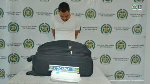 En la fotografía aparece de pie, con los brazos atrás y la cabeza inclinada hacia adelante Jhon Wainer Caicedo Hernández. Viste con una camiseta de color blanca y mangas cortas. Delante de él hay una superficie con tela de color blanco y sobre ella se aprecia una maleta de color negro y una prenda blanca. Sobre la prenda blanca hay una regleta con el escudo de la Policía Nacional y la palabra ‘Cocaína’. Detrás del imputado hay un pendón institucional de la Policía.