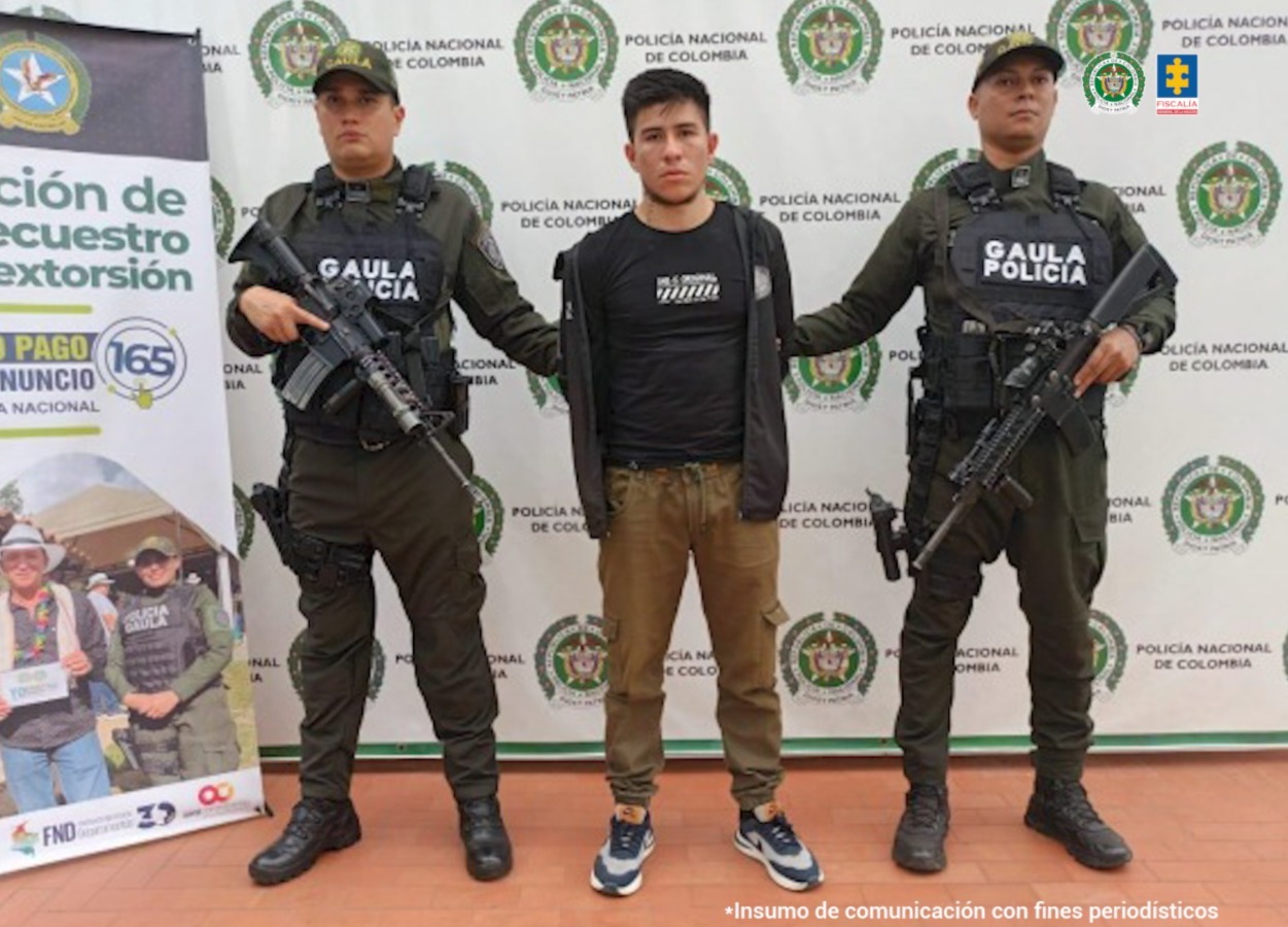 El capturado se encuentra de frente y custodiado por dos uniformados del Gaula de la Policía Nacional. En la foto se ve de fondo un backing de la Policía Nacional.