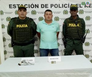 capturado de frente esposado custodiado por policías. Detrás banner de la Policía Nacional.