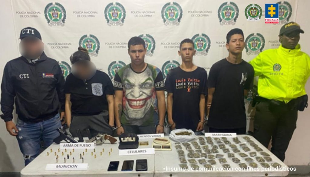 En la fotografía se aprecian de pie y esposados entre sí a los tres imputados. Al extremo derecho de ellos hay un servidor del CTI de la Fiscalía y a la izquierda un uniformado de la Policía Nacional. Delante de ellos hay una superficie y sobre ella hay armas de fuego, proveedores, munición, celulares y gran cantidad de bolsas plásticas transparentes con contenido vegetal. Detrás de ellos hay un pendón institucional de la Policía.