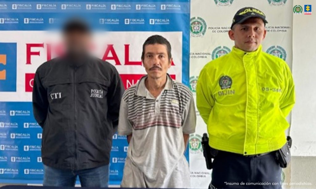 Se observa al capturado de frente. A un costado un funcionario del CTI y al otro uno de Policía Nacional. Atrás un pendón de la Fiscalía General de la Nación y otro de Policía nacional
