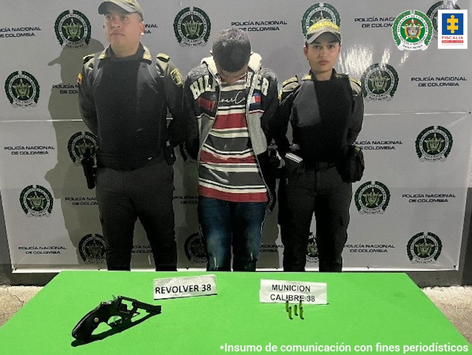 En la foto aparece Jhonier Orlando Herrera Castañeda con los brazos atrás; viste chaqueta y camiseta con rayas rojas, blancas y oscuras y jean azul. A lado y lado de él hay dos uniformados de la Policía Nacional. Delante de ellos una superficie verde con un revólver y 5 cartuchos con los respectivos letreros que dicen ‘Revólver 38’ y ‘munición calibre 38. Detrás de ellos hay un pendón institucional de la Policía Nacional.