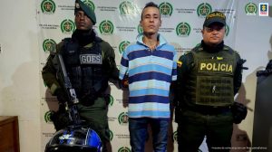 En la foto aparece de pie y con los brazos atrás, Jhonnattan Castillo Villada. Viste camisa de manga corta y cuello, con franjas gruesas horizontales de diferentes tonalidades de azul; así como pantalón azul oscuro. Al lado y lado de él hay uniformados de la Policía Nacional. Detrás de ellos hay un pendón institucional de la Policía.