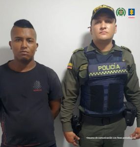 En la fotografía se aprecia de pie  y con los brazos atrás a Alejandro Urbano Martínez. Viste camiseta de manga corta de color negro y pantalón de color negro. A su izquierda hay un uniformado de la Policía Nacional.