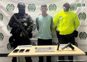 En la fotografía aparece de pie y con los brazos atrás Joan Esteban Rivera De La Cruz, alias de Miami. Viste camiseta de manga corta de color verde claro y pantalón negro. A lado y lado de él hay uniformados de la Policía Nacional. Delante de ellos hay una mesa y sobre ella se parecían tres teléfonos celulares, un proveedor, munición y un arma de fuego. Asimismo, dos letreros que dicen: ´Captura por orden judicial’, ‘Celular incautado’, ‘Arma de fuego’.