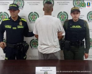 En la fotografía se aprecia de espaldas a la cámara, a Johan Alexis Montenegro Marín. Viste camiseta de manga corta de color blanca y pantalón gris. A lado y lado de él hay uniformados de la Policía Nacional. Delante de ellos hay una superficie marrón y sobre ella un letrero que dice: ‘Violencia intrafamiliar’. Detrás de ellos hay un pendón institucional de la Policía.