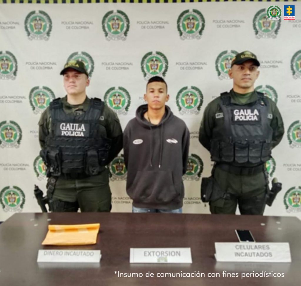 En la fotografía se aprecia de pie y con los brazos atrás a Johan Andrés Otálvaro Soto. Viste con buso de color negro y Jean azul. A sus extremos hay uniformados de la Policía Nacional. delante de ellos hay una superficie de color marrón y sobre ella hay tres letreros que dicen: ‘Dinero incautado’ y detrás de él hay un sobre amarillo; asimismo, hay otro letrero que dice: ‘Extorsión’, y otro que dice: ‘Celulares incautados’, detrás de este último hay un teléfono móvil. Detrás del capturado y los policías hay un pendón institucional de dicha institución.