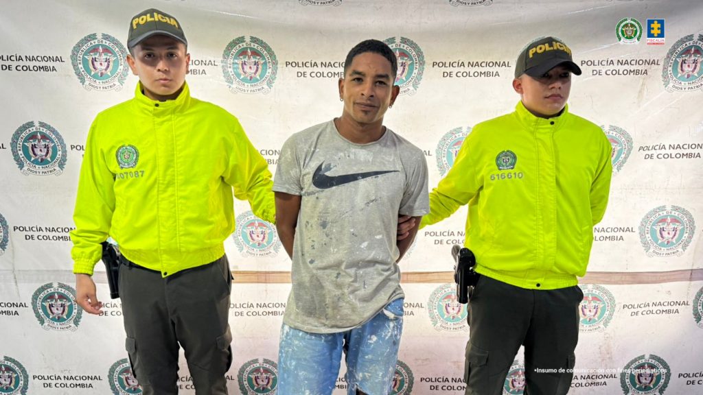 En la fotografía se aprecia de pie y con los brazos atrás a Jhoan Bernardo Quiñónez, alias Chapo. Viste camiseta de color gris con estampado negro a la altura del pecho; así como jean azul desgastado. A sus extremos hay uniformados de la Policía Nacional. Detrás de ellos hay un pendón institucional de la Policía.