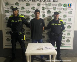 en la imagen se ve una persona detenida bajo custodia de dos integrantes de la Policía Nacional. Frente a él una mesa con municiones incautadas. Detrás suyo un backing institucional.