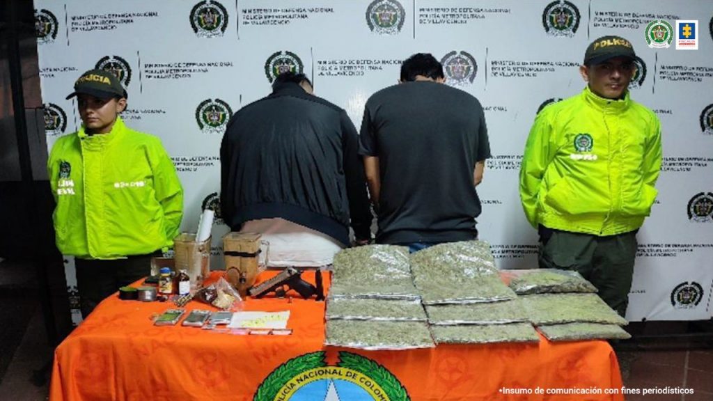 Está el capturado con otra persona de espaldas, custodiados por dos policías, delante de ellos una mesa con el material incautado y detrás banner de la Policía.