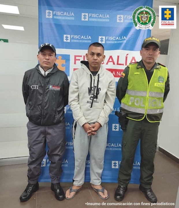 En la foto se ve al detenido esposado, con una sudadera, mirando de frente custodiado por un uniformado de la Policía y un investigador del CTI de la Fiscalía a lado y lado del detenido detrás el banner de Fiscalía.