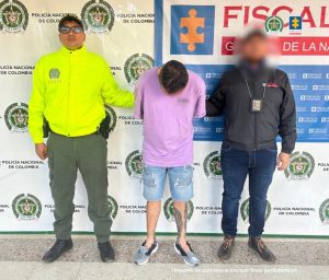 En la imagen se ve al capturado en custodia por un servidor del Cuerpo Técnico de Investigación CTI y un uniformado de la Policía Nacional. El investigado tiene un buzo color lila y una bermuda de jean.
