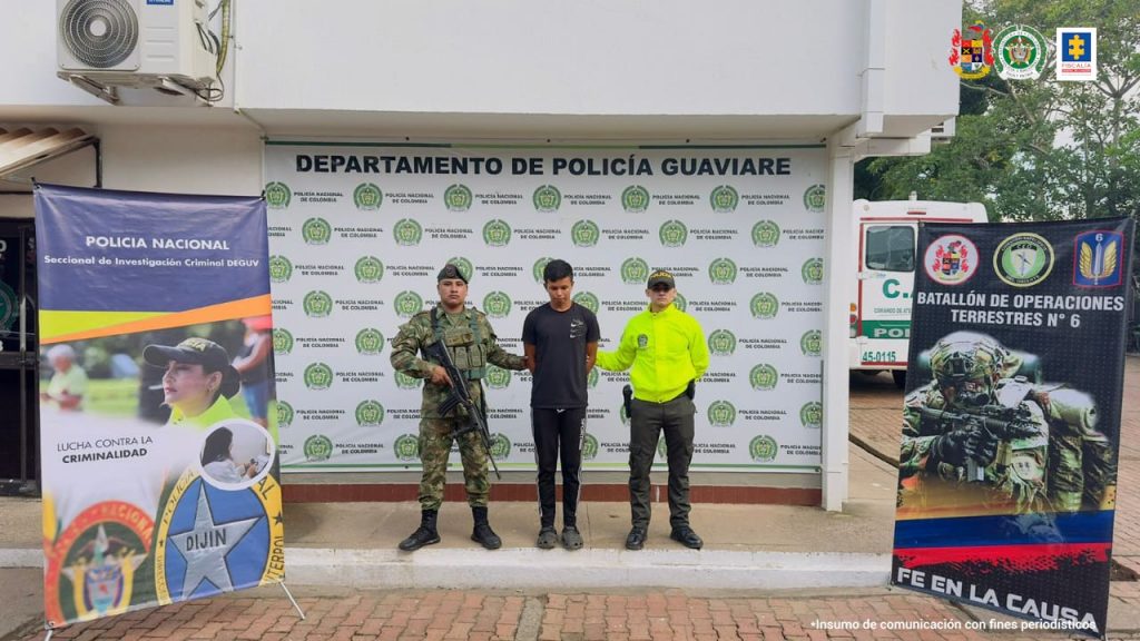 capturado de frentes masno atrás esposado cvustodiado por un militar y un policía. a los lados y dertrás banner de la Policía y del Ejército Nacional.