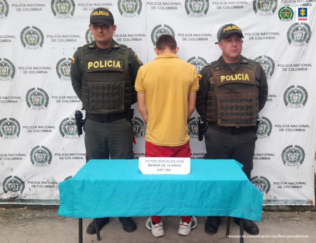 En la foto aparece Jonathan Alberto Velasco Ossa de espaldas, con dos policías a los lados, adelante una mesa con mantel azul con un letrero que dice, actos sexuales con menor de 14 años, y un pendón de la Policía Nacional atrás.