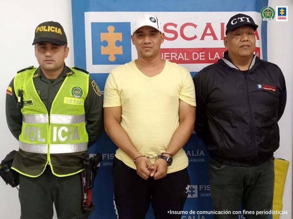 En la foto aparece de pies Jonathan Fernando Vasco Becerra, con los brazos adelante y esposado. Vestido con camiseta color crema, gorra blanca, pantalón y zapatos negros. Al lado derecho de él hay un uniformado de la Policía Nacional y a su izquierda un servidor del CTI de la Fiscalía. Detrás de ellos hay un pendón institucional de la Fiscalía.