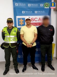 En la fotografía se aprecia de pie, con los brazos adelante y esposado a Jonathan Fernando Vasco Becerra. Viste con gorra blanca, camiseta de manga corta, de color amarillo; pantalón de sudadera negro y zapatillas negras con blanco. Al lado izquierdo de él hay un servidor del CTI de la Fiscalía y al lado derecho un uniformado de la Policía Nacional. Detrás de ellos hay un pendón institucional de la Fiscalía.