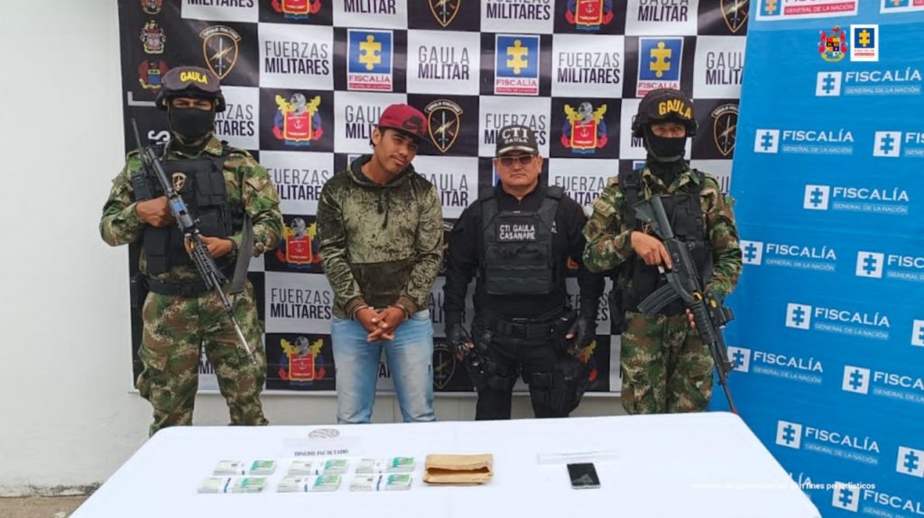 capturado con gorra y esposado manos adelanre, custodiado por personal del CTI de la Fiscalía y militares del Ejército Nacional. Delante mesa con material incautado al detenido, detrás banner de fiscalía y ejército.