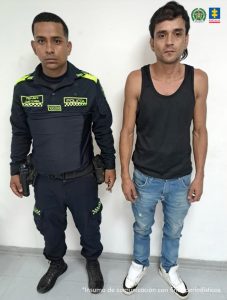 En la fotografía se aprecia de pie a Jordan David Moreno. Viste camiseta esqueleto de color negro, jean azul claro desgastado y zapatillas blancas. Al lado derecho de él hay un uniformado de la Policía Nacional.