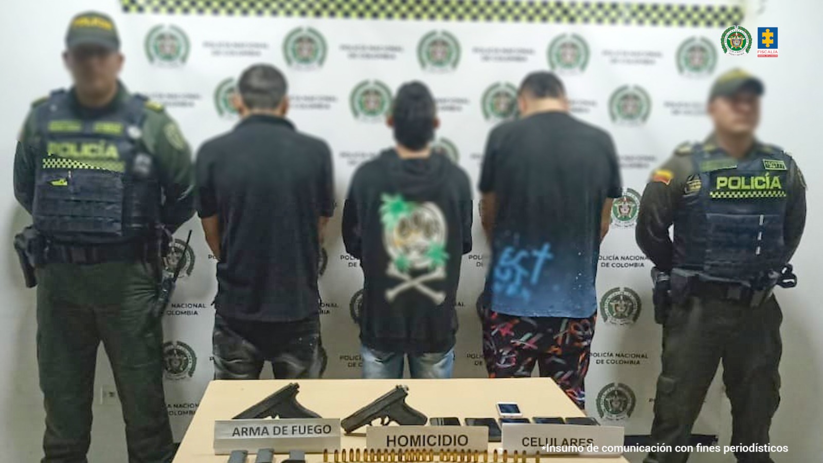 Se observan los tres capturados en fila y de espaldas. A cada lado de la fila, un funcionario de Policía Nacional custodiándolos. Atrás un pendón de Policía nacional. Adelante, una mesa con el material incautado