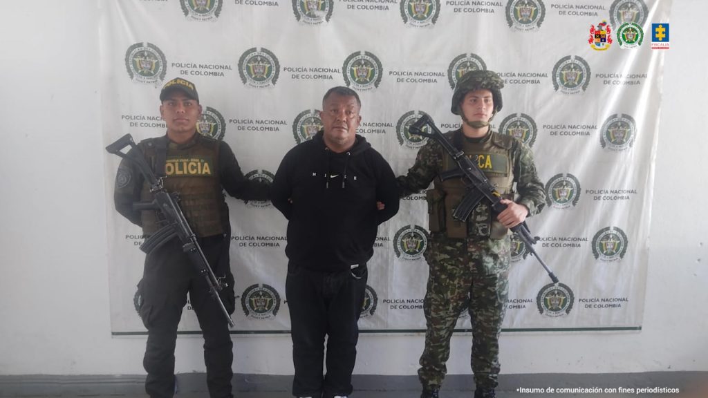 En la fotografía se aprecia de pie y con los brazos atrás a Jorge Arturo Salazar Caicedo, alias ‘El Chamán’. Viste conjuto de sudadera completo de color negro. A sus extremos hay uniformados de la Policía y Ejército. Detrás de ellos hay un pendón institucional de la Policía.