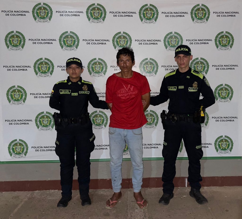 capturado de frente custodiado por dos uniformados. Banner de la Policía Nacional.