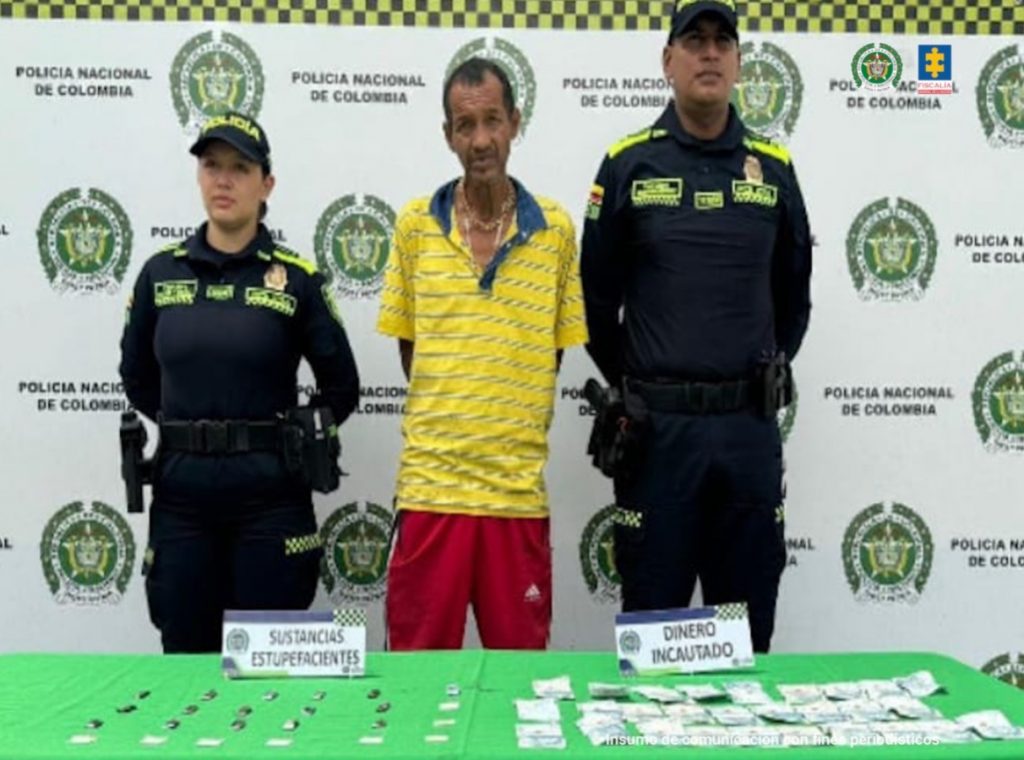 En la fotografía se aprecia de pie y con los brazos atrás a Jorge Iván Guarnizo Cardona. Viste camisa amarilla con rayas azules horizontales, delgadas y pantaloneta roja. A sus extremos hay uniformados de la Policía Nacional. Delante de ellos hay una superficie con mantel verde y sobre ella hay bolsas plásticas pequeñas y dinero en efectivo. Asimismo, hay dos letreros que dicen: ‘Sustancias estupefacientes’ y ‘Dinero incautado’. Detrás de ellos hay un pendón institucional de la Policía.
