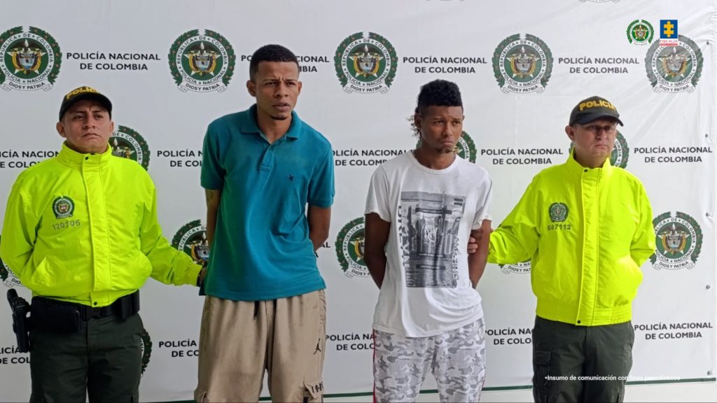 En la fotografía aparecen dos hombres capturados en compañía de dos uniformados de la Policía Nacional. En la parte posterior un banner con logos de la entidad.