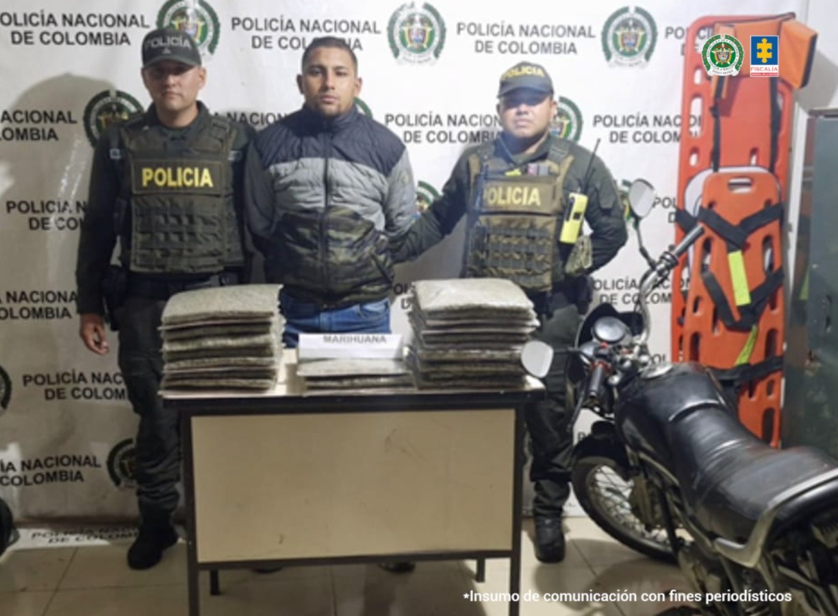En la fotografía se aprecia de pie y con los brazos atrás a Jorge Luis Grisales Sánchez. Viste chaqueta de color gris con negro y jean azul. A sus extremos hay uniformados de la Policía Nacional. Delante de ellos hay una mesa y sobre ella hay varios paquetes plásticos con sustancia vegetal; asimismo, un letrero que dice ‘Marihuna’. Al lado izquierdo del judicializado hay una motocicleta. Detrás de ellos hay un pendón institucional de la Policía.
