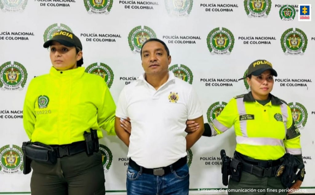 En la fotografía aparece un hombre capturado, acompañado de dos uniformados de la Policía Nacional.   En la parte posterior un banner con logos de la institución.  