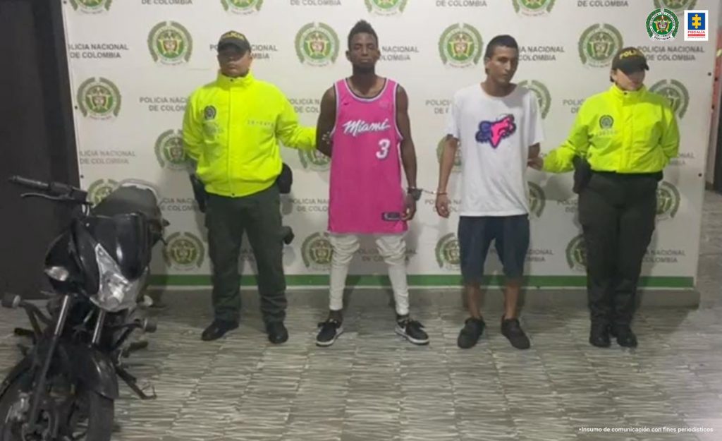 En la foto aparecen de pie José Andrés Grueso Martínez y Dillen David Cruz. Uno viste camiseta esqueleto color fucsia, pantalón blanco y zapatos negros. La otra persona vista camiseta de manga corta blanca con estampado a la altura del pecho, pantaloneta oscura y zapatos negros. A lado y lado de ellos hay uniformados de la Policía Nacional y a la derecha de ellos hay una motocicleta.