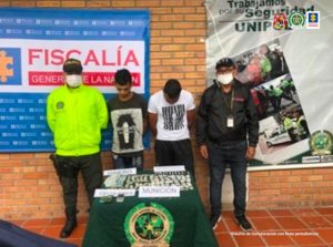 En la fotografía aparecen dos hombres capturados junto a investigadores del CTI y de la Policía Nacional. En frente de los detenidos hay una mesa en la que se observa el material incautado en la diligencia de allanamiento y en la parte posterior se encuentra un banner de la Fiscalía y de la Policía Nacional.