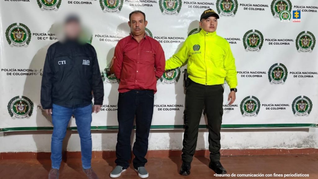 El capturado se encuentra de frente y custodiado por un servidor del Cuerpo Técnico de Investigación CTI y un uniformado de la Policía Nacional. Detrás se observa un backing de la Policía Nacional.