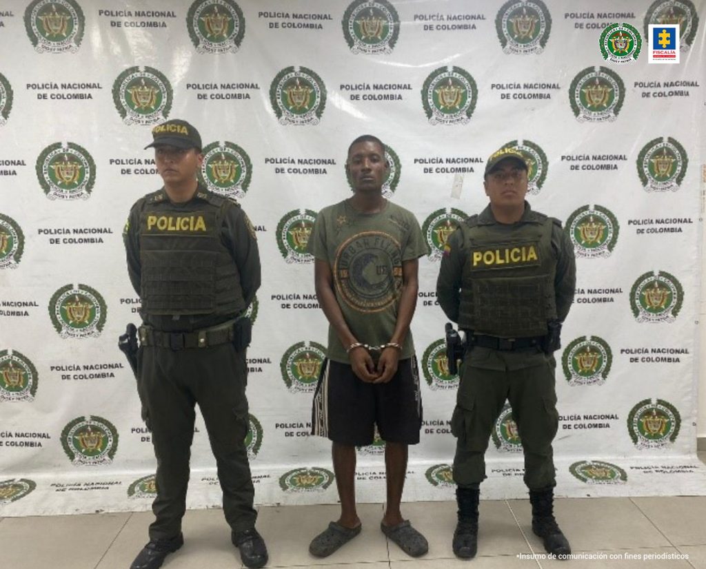 En la foto aparece de pie, con los brazos adelante y esposado, José David Arango Gutiérrez. Viste camisa verde oliva, de manga corta, con estampado circular amarillo; pantaloneta oscura y pantuflas oscuras. Al lado y lado de él hay uniformados de la Policía Nacional. Detrás de ellos hay un pendón interinstitucional de la Policía.