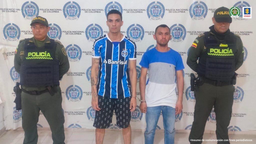 En la fotografía aparecen dos hombres capturados, acompañados de dos uniformados de la Policía Nacional. En la parte posterior un banner con logos de la entidad.