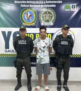 capturado de frente custodiado por dos policías. Detrás banner de Policía Nacional.