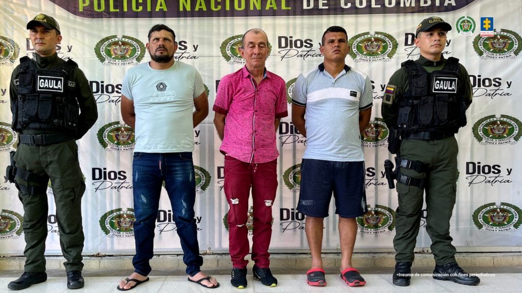 Capturados de frente custodiados por dos policías. Detrás banner de la Policía Nacional.