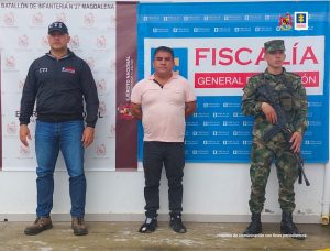 En la fotografía aparece un capturado, junto a personal de Fiscalía General de la Nación y Ejército Nacional. En la parte posterior de la imagen se ven banners de Ejército Nacional y la Fiscalía General de la Nación. 