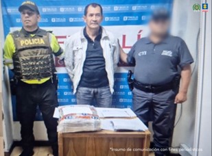 Se visualiza al capturado junto a un uniformado de la Policía Nacional y un investigador del CTI. Frente a ellos una mesa con las sustancias incautadas.  Detrás de ellos el banner que identifica a la Fiscalía General de la Nación.