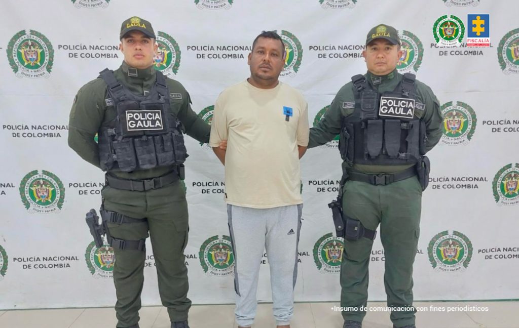 En la imagen aparece una persona de suéter amarillo y sudadera gris, entre dos uniformados de la Policía Nacional.