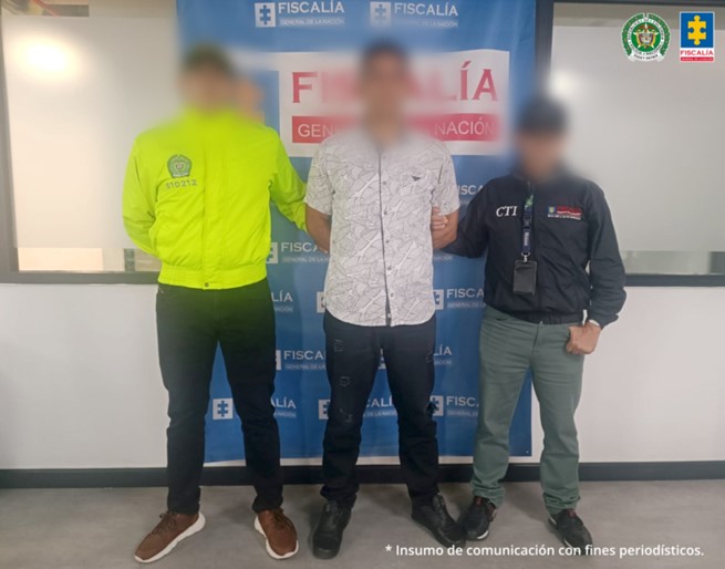 Se visualiza al capturado con el rostro difuminado junto a un uniformado de la Policía Nacional y un investigador del CTI. Detrás de ellos se ubica el banner institucional de la Fiscalía General de la Nación.
