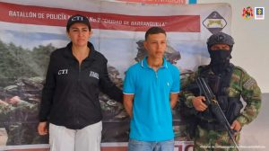 capturado de frente manos atrás custodiado por investigadora del CTI de la Fiscalía y militar de Ejército Nacional. Detrás banner de Fiscalía y Ejército Nacional.