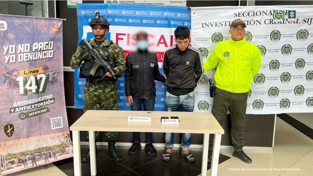 Se visualiza al capturado junto a una investigadora del CTI y uniformados del Ejército y Policía Nacional. En la parte posterior se ubican los banners institucionales de la Fiscalía General de la Nación, del Ejército y la Policía Nacional.