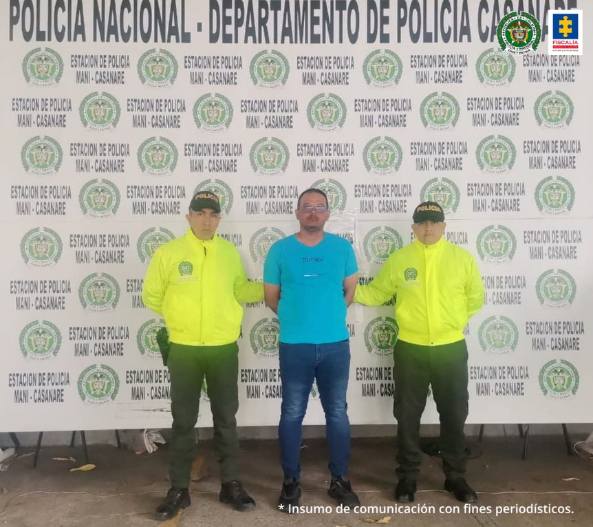 Capturado con gafas y manos esposadas atrás, custodiado por dos policía. Detrás banner de Policía