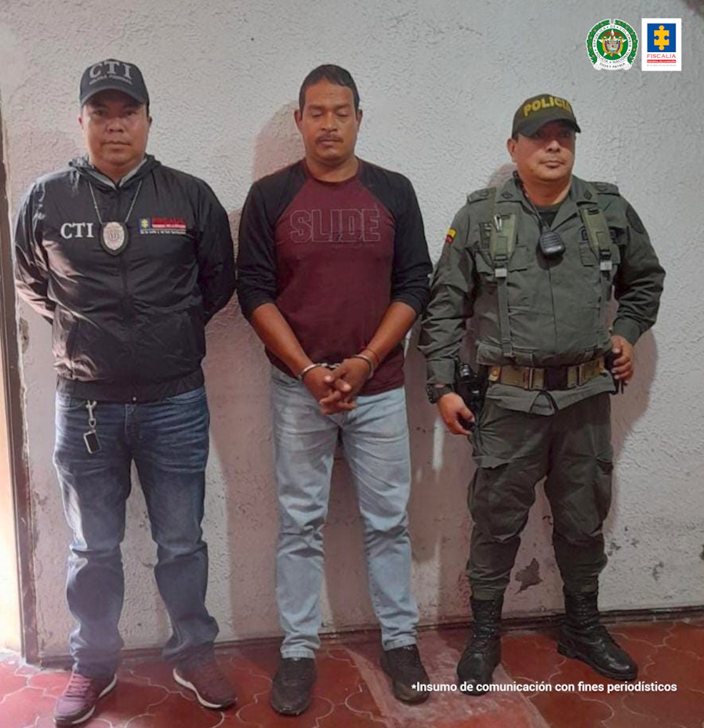 En la foto aparece el judicializado vestido con buso vinotinto de mangas negras, jean azul claro y zapatos negros; esposado con sus manos al frente. Al lado derecho aparece un servidor del CTI de la Fiscalía y a su izquierda uno de la Policía Nacional.